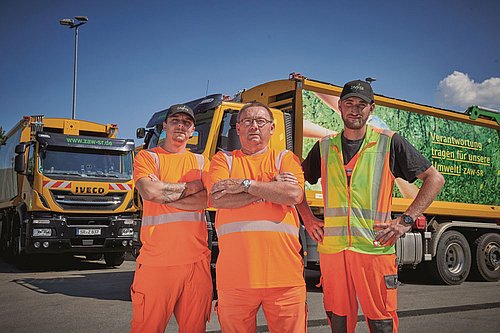 Drei Mitarbeiter der Müllabfuhr mit Entsorgungsfahrzeugen im Hintergrund