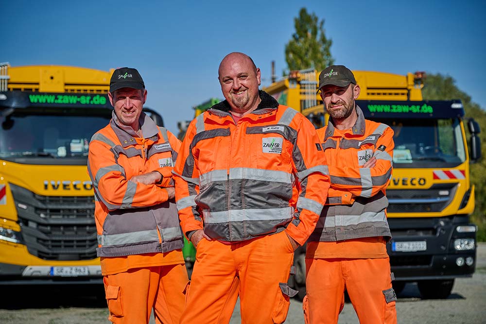 Drei Müllwerker mit gelben Müllabfuhr-Fahrzeugen im Hintergrund